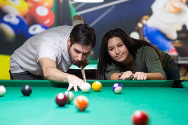 A guy and a young and beautiful woman are playing billiards in a club. They are having a good time. 
