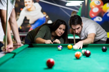 A guy and a young and beautiful woman are playing billiards in a club. They are having a good time. 