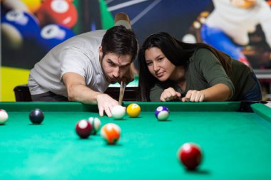 A guy and a young and beautiful woman are playing billiards in a club. They are having a good time. 