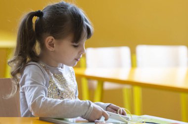 6-7 yaşlarında bir kız okul bankında oturur ve eğitim kitabıyla eğlenir. 