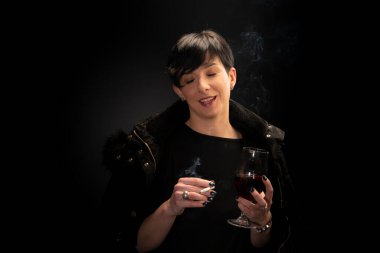 Genç bir kadın şarap içiyor ve siyah arka planda sigara içiyor.