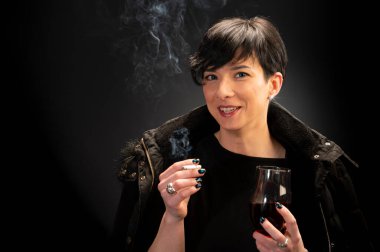 Genç bir kadın şarap içiyor ve siyah arka planda sigara içiyor.