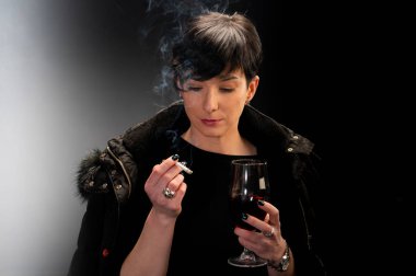 Genç bir kadın şarap içiyor ve siyah arka planda sigara içiyor.