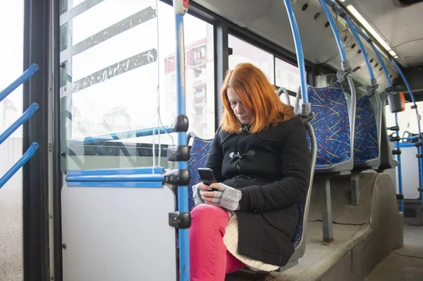 A middle-aged woman with red hair is sitting on a seat and having fun with her phone while riding a bus