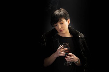 Genç bir kadın şarap içiyor ve siyah arka planda sigara içiyor.