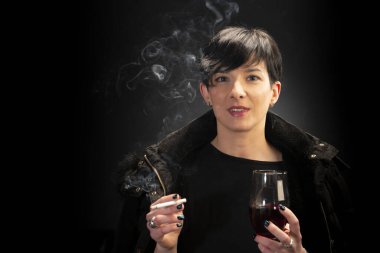 Genç bir kadın şarap içiyor ve siyah arka planda sigara içiyor.