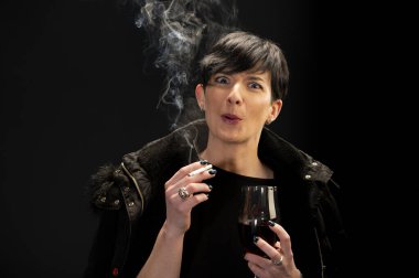 Genç bir kadın şarap içiyor ve siyah arka planda sigara içiyor.