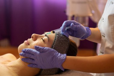Güzellik kliniğinde gençleştirici yüz enjeksiyonları yaptıran güzel bir genç kadın. Kozmetik uzmanı bir müşterinin derisine yüz doldurucu enjekte ediyor.