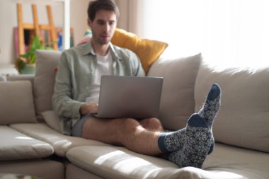 Ev ortamında rahat bir kanepede çalışan genç bir adam dizüstü bilgisayar kullanıyor. Uzak çalışma, bağlantı, serbest meslek, serbest yaşam tarzı konsepti