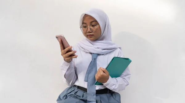 印度尼西亚高中女生身穿白色和灰色制服 手持手机和笔记本坐在那里 她的眼睛看着手机 — 图库照片