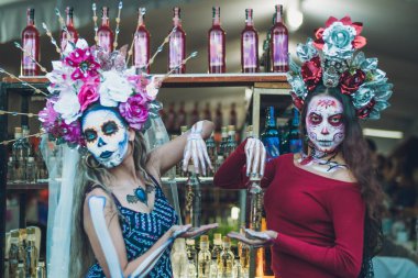 Meksikalı bir kadın, Ölüler Günü şenliği için kedi gibi giyinmiş ve karakterize edilmişti. Elinde bir şişe mezcal taşıyordu. Arkasında mezcal şişelerden oluşan bir arka plan vardı..
