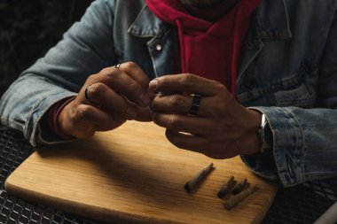 Elinde farklı boyutlarda marihuana sigaraları olan bir adam ahşap bir tahtanın üzerinde, etrafta marihuana kalıntılarıyla....