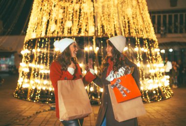 Arkadaşlar ışıkları izlerken eğleniyorlar, kışlık kıyafetlerini giyiyorlar Noel alışverişi yapıyorlar, çantaları hediyelerle dolu, arka plandaki ışıklar kocaman bir Noel ağacı..