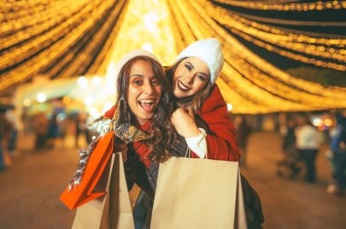 Arkadaşlar ışıkları izlerken eğleniyorlar, kışlık kıyafetlerini giyiyorlar Noel alışverişi yapıyorlar, çantaları hediyelerle dolu, arka plandaki ışıklar kocaman bir Noel ağacı..