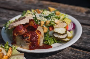 Erimiş peynirli hamburger, pastırma ve biftek, yanında salata ve turşu, tipik Amerikan yemeği, kırsal bir ahşap masada beyaz bir tabakta servis edilir..