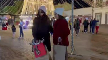 Arkadaşlar ışıkları izlerken eğleniyorlar, kışlık kıyafetlerini giyiyorlar Noel alışverişi yapıyorlar, çantaları hediyelerle dolu, arka plandaki ışıklar kocaman bir Noel ağacı..