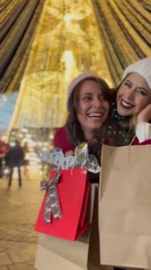 Arkadaşlar ışıkları izlerken eğleniyorlar, kışlık kıyafetlerini giyiyorlar Noel alışverişi yapıyorlar, çantaları hediyelerle dolu, arka plandaki ışıklar kocaman bir Noel ağacı..
