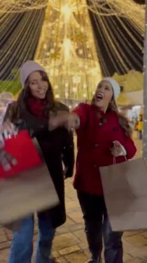 Arkadaşlar ışıkları izlerken eğleniyorlar, kışlık kıyafetlerini giyiyorlar Noel alışverişi yapıyorlar, çantaları hediyelerle dolu, arka plandaki ışıklar kocaman bir Noel ağacı..