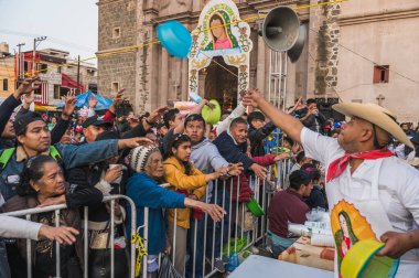 Geleneksel kıyafetler giyen yerli bir adam, Guadalupe Bakiresi kutlamasına katılanlara hediyeler veriyor, arkalarında da kilise var..