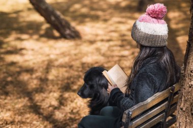 Oryantal özellikleri olan güzel beyaz kız parkta kitap okuyor, köpeğiyle bankta oturuyor ve dallardan gün batımı ışığı süzülüyor..