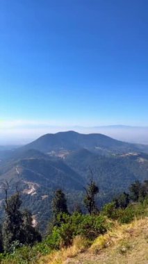 Bir dağın tepesinden gelen dikey 4k video ile ağaçlı, vadili ve arka planda mavi gökyüzü ve sisli arasında kaybolan küçük dağları görebilirsiniz..