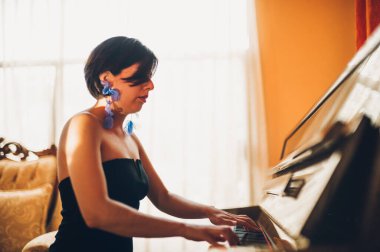 Güzel piyanist kız ahşap dik piyanosunu çalıyor, siyah ve zarif giyinmiş, mavi çiçekli küpeleri var..