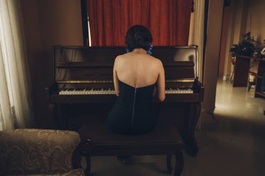 Güzel piyanist kız ahşap dik piyanosunu çalıyor, siyah ve zarif giyinmiş, mavi çiçekli küpeleri var..