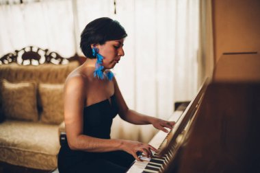 Güzel piyanist kız ahşap dik piyanosunu çalıyor, siyah ve zarif giyinmiş, mavi çiçekli küpeleri var..