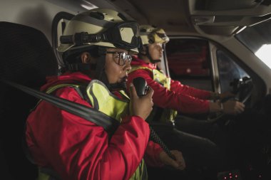 Bir çift Latin erkek ve kadın sağlık görevlisi ambulansın içinde oturur, adam araba kullanır, kadın da telsizden konuşur, arama ihtimaline karşı hayat kurtarmaya hazırdır..