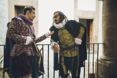 Mor ve yeşil giyinmiş yakışıklı adamlar, 1600 'lü yıllardan kalma giysiler, eski bir malikanenin ikinci katındaki antika korkuluklarda dikilip sohbet ediyorlar..