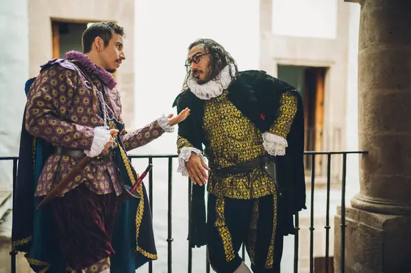 Mor ve yeşil giyinmiş yakışıklı adamlar, 1600 'lü yıllardan kalma giysiler, eski bir malikanenin ikinci katındaki antika korkuluklarda dikilip sohbet ediyorlar..