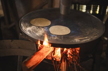 Beyaz işlemeli bluz ve mavi süveter giyen Meksikalı bir kadın geleneksel Meksika yemeği olan tortilla yapmak için komalın üzerine yassı hamuru yerleştiriyor..