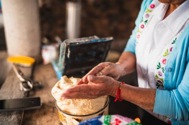 Beyaz işlemeli bluz ve mavi kazak giyen Meksikalı bir kadın geleneksel Meksika yemeği olan tortilla yapmak için hamuru elleriyle hazırlıyor..