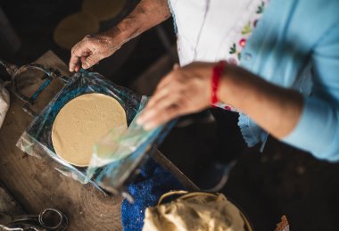 Beyaz işlemeli bluz ve mavi süveter giyen Meksikalı bir kadın geleneksel Meksika yemeği olan tortilla yapmak için tortilla makinesine hamur sıkıştırıyor..