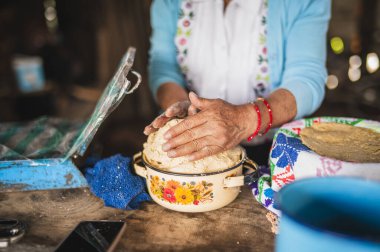 Beyaz işlemeli bluz ve mavi kazak giyen Meksikalı bir kadın geleneksel Meksika yemeği olan tortilla yapmak için hamuru elleriyle hazırlıyor..