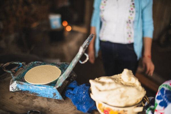 An indigenous Mexican woman wearing a white embroidered blouse and blue sweater, pressing dough into a tortilla press to make tortillas, a traditional Mexican dish.
