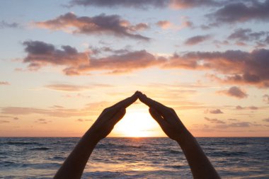 Eller bir çatı şeklinde katlanmış, güneşin üzerinde bir ev. Siluet. Gün ışığı, gün batımı ya da şafak. Çevre koruma, hayat sigortası, refah konsepti. Deniz dalgaları, renkli gökyüzü.