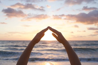 Eller bir çatı şeklinde katlanmış, güneşin üzerinde bir ev. Siluet. Gün ışığı, gün batımı ya da şafak. Çevre koruma, hayat sigortası, refah konsepti. Deniz dalgaları, renkli gökyüzü.