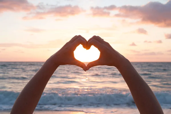 Kalp şeklinde kadın elleri, gün batımında güneş ışığı, ya da arka planda şafak vakti gökyüzü. Kalp şeklinde eller, tatil, seyahat, aşk konsepti. Deniz ya da okyanus dalgaları, renkli gökyüzü.