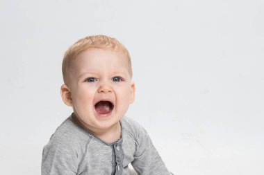 Beyaz stüdyo arka planında gri elbiseli, ağzı açık, neşeli bir bebek. Sağlıklı çocuk kavramı, 0 yaşından 1 yaşına kadar yeni yürümeye başlayan çocuk. Uzayı kopyala Göz teması. Beyaz çocuk, sarı saç, mavi göz.