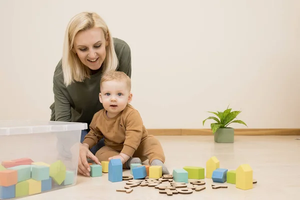 Mom plays with baby at home, child builds a tower, colored blocks. Mother and little son. Concept of time together, early development, parenthood, storage toys. Caucasian family. Indoor. Wooden floor.