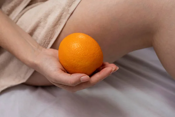 Orange in the hands of a girl, bare legs on the background. Concept of cosmetology, diet, vitamin C. body care, skin, cellulite, fetal size. Day light.