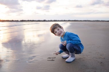 Child on the beach, draws on the sand near the sea. Boy in a blue sweatshirt and rubber boots. Autumn, beautiful sky, clouds reflected in the water. Sunset. Concept son, children, happiness, childhood