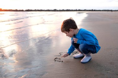 Child on the beach, draws on the sand near the sea. Boy in a blue sweatshirt and rubber boots. Autumn, beautiful sky, clouds reflected in the water. Sunset. Concept son, children, happiness, childhood