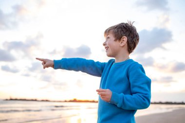 Şaşkın çocuk arka planda bulutlarla parmağını, denizini ve gökyüzünü işaret ederek elini ileri uzattı. Kumsal, gün batımı. Seyahat, tatil, çocukluk kavramı. Beyaz çocuk, 6 yaşında..