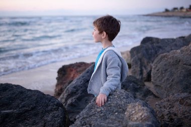 Surprised child on the seashore standing between large stones.  Autumn, storm on the sea.  The concept of childhood, emotions, nature, impressions, travel. 6 years old boy in profile.