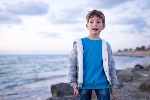 stock image Handsome caucasian boy walks near the sea. Child in casual autumn clothes, gray cardigan, blue sweatshirt. Rocky beach, waves. Concept of travel, nature, pastime, outdoor hiking, tourism. Sunset. 