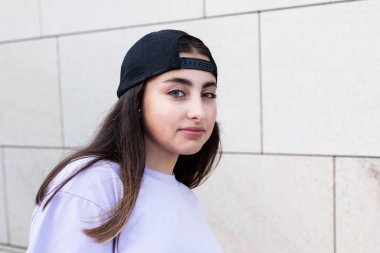 Pozitif, kendine güvenen genç bir kızın portresi. Dışarı çıkın. Havalı genç bir kadın, sokak tarzı. Güneşli bir gün. Doğal güzellik, ten, yaşam tarzı konsepti. Kafkasyalı esmer. Arka plandaki binanın duvarı..