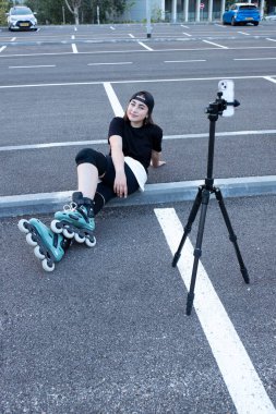 Boş bir otoparkta oturup selfie çeken bir kız. Ergen, ayakları üzerinde paten kayıyor, kendini telefonun ön kamerasında tripodun üzerinde çekiyor. Video günlüğü, hobi, içerik, vlog, blog yazarı kavramı.