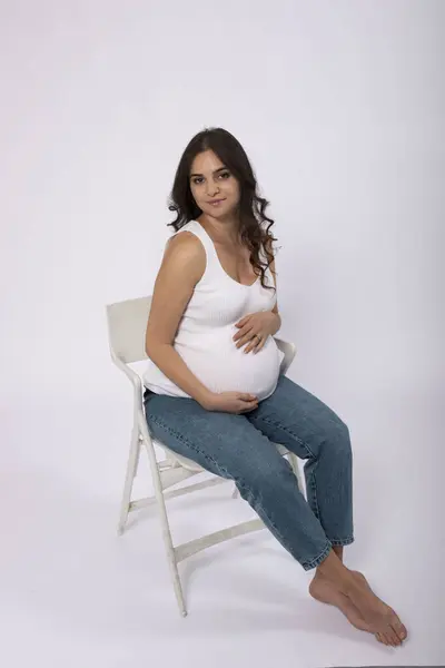 Pregnant brunette woman with long hair. Expectant mother gently touches her tummy while sitting on a chair. Studio light, white background. Concept of waiting for a baby, healthy pregnancy, 9 months .